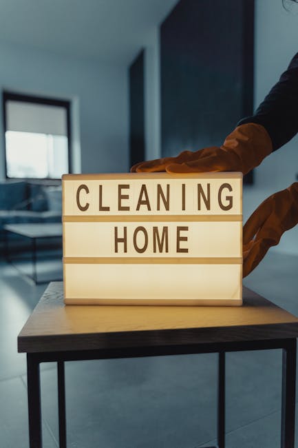 A lightbox sign displaying the words 'CLEANING HOME' on a wooden table in a modern, well-lit room with large windows and a dark wall in the background. The sign is illuminated, emphasizing clean and p