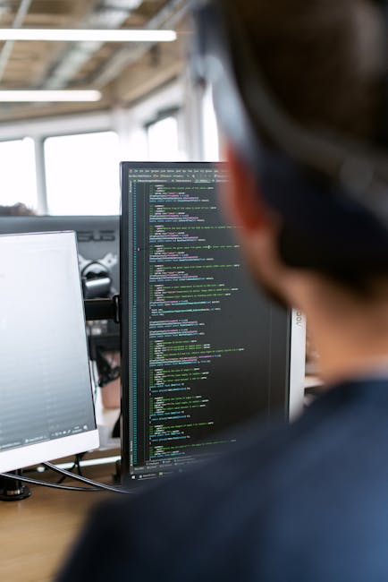 A person working on a laptop with a dark-themed code editor open on the screen, displaying lines of green, blue, and white text and symbols. The individual uses their right hand to operate the trackpa